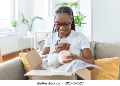 Young Satisfied Happy African Girl Woman Lady Shopaholic Customer Sit On Sofa Unpack Parcel Delivery Box, Online Shopping Shipment Concept. Taking Photos Of Product To Post On Social Media