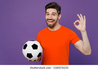 Young Satisfied Happy Fun Fan Man He Wears Orange T-shirt Cheer Up Support Football Sport Team Hold In Hand Soccer Ball Watch Tv Live Stream Showing Okay Ok Gesture Isolated On Plain Purple Background