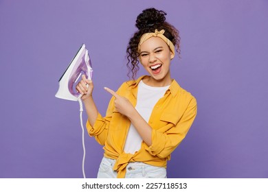 Young satisfied happy fun housekeeper woman wear yellow shirt tidy up hold in hands point index finger on iron isolated on plain pastel light purple background studio. Housework housekeeping concept