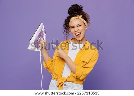 Young satisfied happy fun housekeeper woman wear yellow shirt tidy up hold in hands point index finger on iron isolated on plain pastel light purple background studio. Housework housekeeping concept