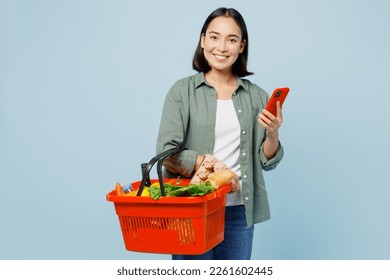 Young satisfied happy fun woman wear casual clothes hold red basket with food products use mobile cell phone isolated on plain blue background studio portrait. Delivery service from shop or restaurant - Powered by Shutterstock