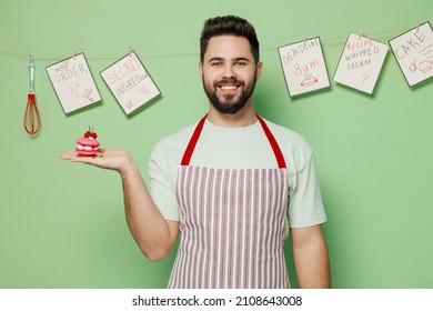 Young Satisfied Happy Male Chef Confectioner Baker Man In Striped Apron Holding Pink Little Cake Muffin Macaroon Isolated On Plain Pastel Light Green Background Studio Portrait. Cooking Food Concept