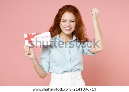 Young satisfied happy student redhead woman 20s wearing blue shirt holding gift voucher flyer mock up do winner gesture clench fist celebrating isolated on pastel pink color background studio portrait