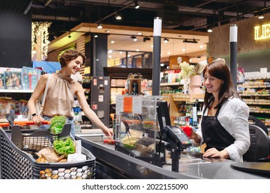 Young satisfied happy woman 20s wear casual clothes shopping at supermaket with grocery cart stand at store checkout pays for groceries cashier inside hypermarket Purchasing gastronomy food concept.