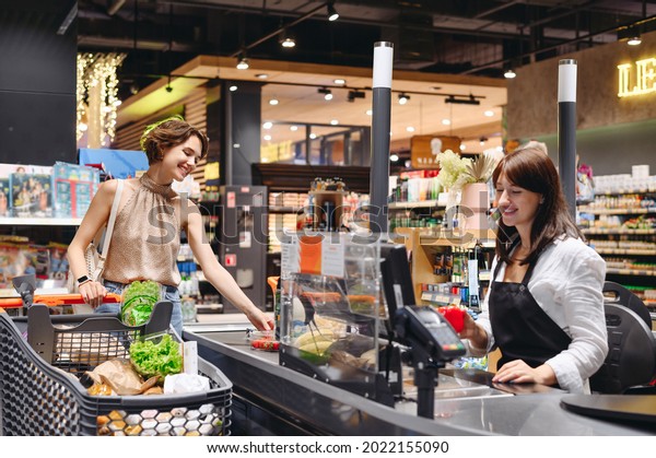 Young satisfied happy woman 20s wear casual\
clothes shopping at supermaket with grocery cart stand at store\
checkout pays for groceries cashier inside hypermarket Purchasing\
gastronomy food concept.