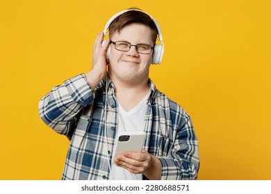 Young satisfied man with down syndrome wear glasses casual clothes headphones listen to music use mobile cell phone isolated on pastel plain yellow color background. Genetic disease world day concept
