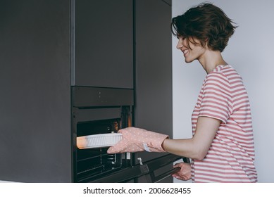 Young Satisfied Side View Housewife Woman In Casual Clothes Striped T-shirt Puts Casserole In Baking Dish In Oven Cooking Food In Light Kitchen At Home Alone. Healthy Diet Bakery Lifestyle Concept.