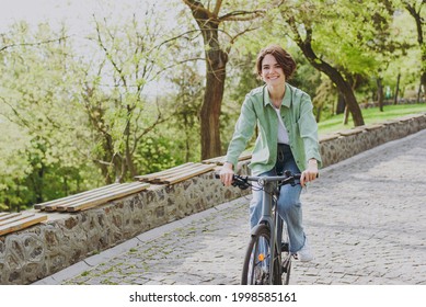 Young satisfied smiling happy woman 20s wear casual green jacket jeans riding bicycle bike on sidewalk in city spring park outdoors, look camera. People active urban healthy lifestyle cycling concept. - Shutterstock ID 1998585161