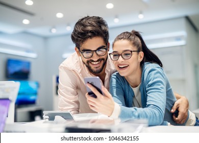 Young satisfied stylish charming couple testing the new model of a mobile from the desk in the tech store. - Shutterstock ID 1046342155