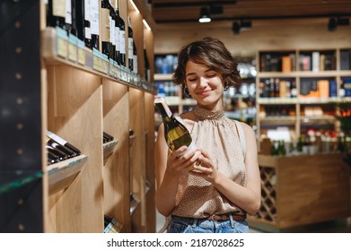 Young Satisfied Woman 20s In Casual Clothes Shopping At Supermaket Grocery Store Buy Choosing White Wine Alcohol Hold Bottle Prop Up Chin Inside Hypermarket. People Purchasing Gastronomy Food Concept