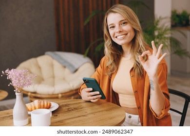 Young satisfied woman wear orange shirt hold use mobile cell phone show ok okay gesture sitting alone at table in coffee shop cafe restaurant indoors work or study. Freelance office business concept - Powered by Shutterstock