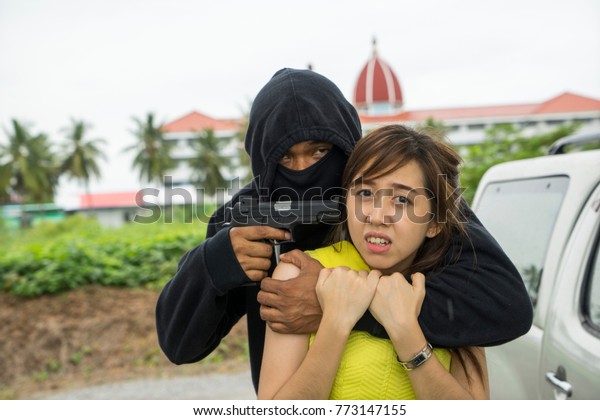 Young scared woman victim locked by\
robber near her car in day time Gangster criminal cover face with\
black mask pointing gun at hostage. Criminal, kidnap, terrorist,\
property & life Insurance\
concept