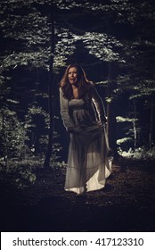 Young Scared Woman Walking Through The Forest At Night In White Dress And Screaming. Horror Scene.