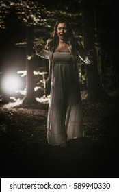 Young Scared Woman Walking Through The Forest At Night In White Dress And Screaming. Horror Scene.