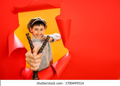 A young school boy in a retro cap and knit sweatshirt is aiming from a wooden slingshot to the target. The child looks into a hole with curls in a red studio background. Copy space.