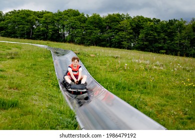 Young School Kid Boy Having Fun Riding Summer Toboggan Run Sled Down A Hill In Hoherodskopf, Germany. Active Child With Medical Mask Making Funny Activity Otudoors. Family Leisure With Kids.