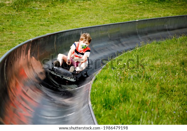 Young school kid boy having fun riding summer\
toboggan run sled down a hill in Hoherodskopf, Germany. Active\
child with medical mask making funny activity otudoors. Family\
leisure with kids.