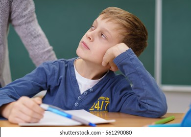 Young Schoolboy Listening To His Teacher As She Leans Over Him At His Desk From Behind In The Classroom, Close Up On The Child