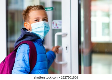 Young Schoolboy Wearing Protective Mask Is Trying To Open The School Door. Behind The Backpack Schoolboy Look At Camera