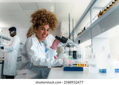 Young scientist smiles while using a microscope in a modern, bright laboratory, wearing a lab coat and pink gloves, with colleagues working in the background, conducting experiments and research