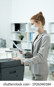 Young Secretary In Grey Suit And Protective Mask Looking Through One Of Papers While Standing By Xerox Machine In Office Environment