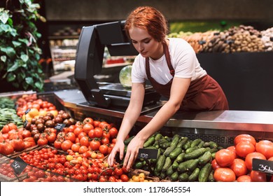 Young seller in apron standing behind counter with vegetables thoughtfully working in modern supermarket