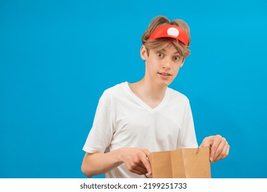 Young Seller In Casual White T-shirt With Paper Bag Isolated On Blue Background Studio Portrait. People Lifestyle Concept. Teen Holding Package Bag With Purchases