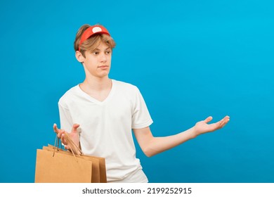 Young Seller In Casual White T-shirt With Paper Bag Isolated On Blue Background Studio Portrait. People Lifestyle Concept. Teen Holding Package Bag With Purchases