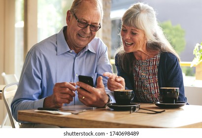 Young senior couple sitting at table in cafe and looking at mobile phone and smiling. Retired man and woman in coffee shop using smart phone. - Powered by Shutterstock