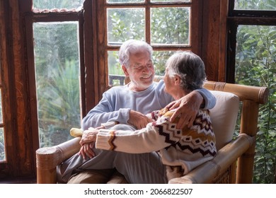 Young Senior Happy Couple Sitting Together On A Bamboo Chair At Home In Wooden Chalet Enjoying Winter Or Autumn Season With Love And Friendship. Old Mature People Lifestyle