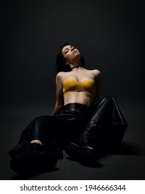 Young sensual languid brunette woman in leather pants, brutal choes and bra sits on floor with head thrown back and eyes closed over black background. Fashion, stylish casual look for woman concept