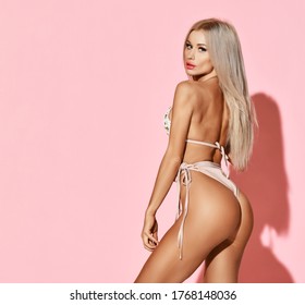 Young sensual slim tanned blond woman in sexy pink beach bikini is posing back to us over pink background with free copy space. Hot summer beach swimsuits and looks for women
