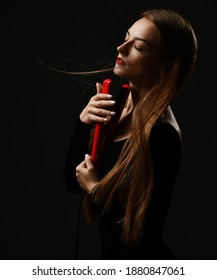 Young sensual woman with long silky fluttering in the wind hair stands with eyes closed holding red hair straightener at face over dark background. Haircare, beauty, wellness concept