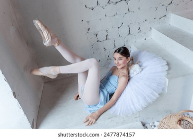 Young serene ballerina posing with white tutu lies on steps, pose relaxed yet elegant, against the rustic texture of white brick wall. Elegant long legs of ballet dancer in pointe shoes