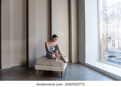 Young serene ballet dancer girl sits on ottoman gracefully securing ribbons of pointe shoes. Sunlight streams through vertical windows, casting soft shadows, enhancing quiet atmosphere of dance studio - Powered by Shutterstock