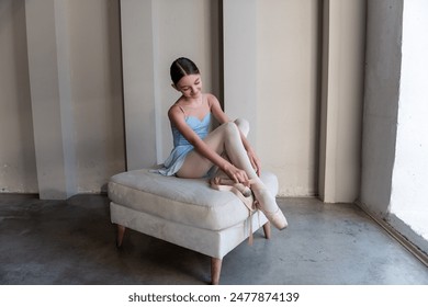 Young serene ballet dancer girl sits on ottoman gracefully securing ribbons of pointe shoes. Sunlight streams through vertical windows, casting soft shadows, enhancing quiet atmosphere of dance studio - Powered by Shutterstock