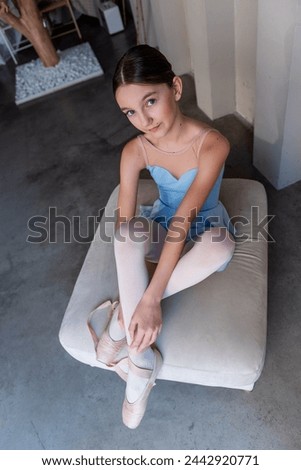 Young serene ballet dancer girl sits on ottoman gracefully securing ribbons of pointe shoes. Sunlight streams through vertical windows, casting soft shadows, enhancing quiet atmosphere of dance studio