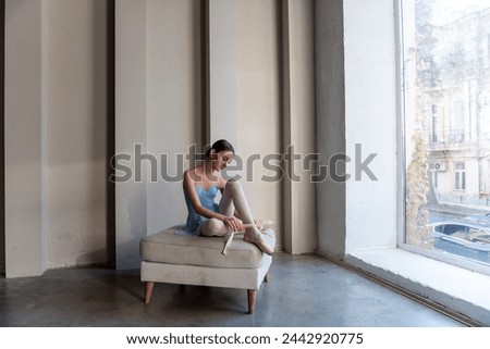 Young serene ballet dancer girl sits on ottoman gracefully securing ribbons of pointe shoes. Sunlight streams through vertical windows, casting soft shadows, enhancing quiet atmosphere of dance studio