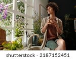 Young serene brunette with cup of coffee looking through window surrounded by plants while sitting by sink in the kitchen