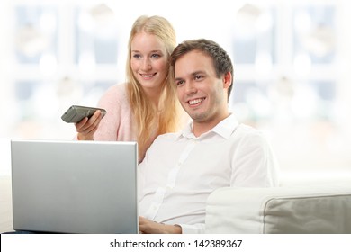 Young serene couple laughing together in their living room at home. Young woman turn on tv while her husband working on laptop.