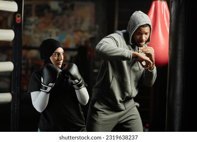 Young serioua Muslim sportswoman in boxing gloves crossing in front of herself and looking at punching bag before hitting it during training - Powered by Shutterstock