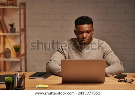 young serious african american freelancer typing on laptop in home office at night, remote work