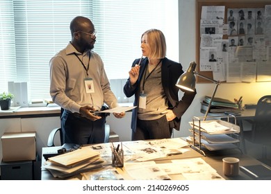 Young Serious African American Male FBI Agent Listening To Mature Female Colleague In Formalwear With Pen By Her Chin