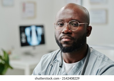 Young Serious African American Male General Practitioner In Blue Uniform And Eyeglasses Sitting By Workplace In Front Of Camera In Hospital