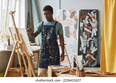 Young serious African American man in denim apron standing in front of canvas on easel and creating new painting at lesiure - Powered by Shutterstock