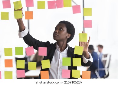Young serious African businesslady jotting on colored adhesive stickers attached on glass wall, write fresh creative start-up ideas, prepare boardroom for briefing. Memory notes, tasks concept