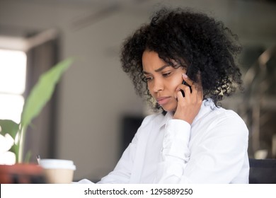 Young Serious African Office Worker Female Sitting At Desk Makes Business Call Listens Client Claim Feels Displeased And Annoyed. Gloomy Mixed Race Employee Using Mobile Phone Solves Problem Distantly