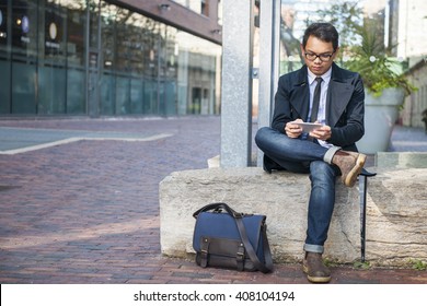 Young Serious Asian Business Man Looking At Mobile Phone Sitting Outside On City Street With Copy Space