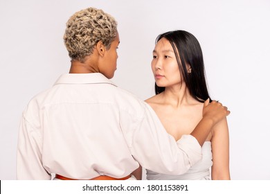 Young serious Asian woman with dark long hair looking at African female standing in front and keeping hand on shoulder of her friend