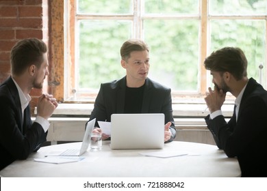 Young Serious Boss In Front Of Laptop Consulting Two Male Employees During Briefing Meeting. Executive Employee Talking About Company Goals To Coworkers. Manager Sharing Important Corporate News. 